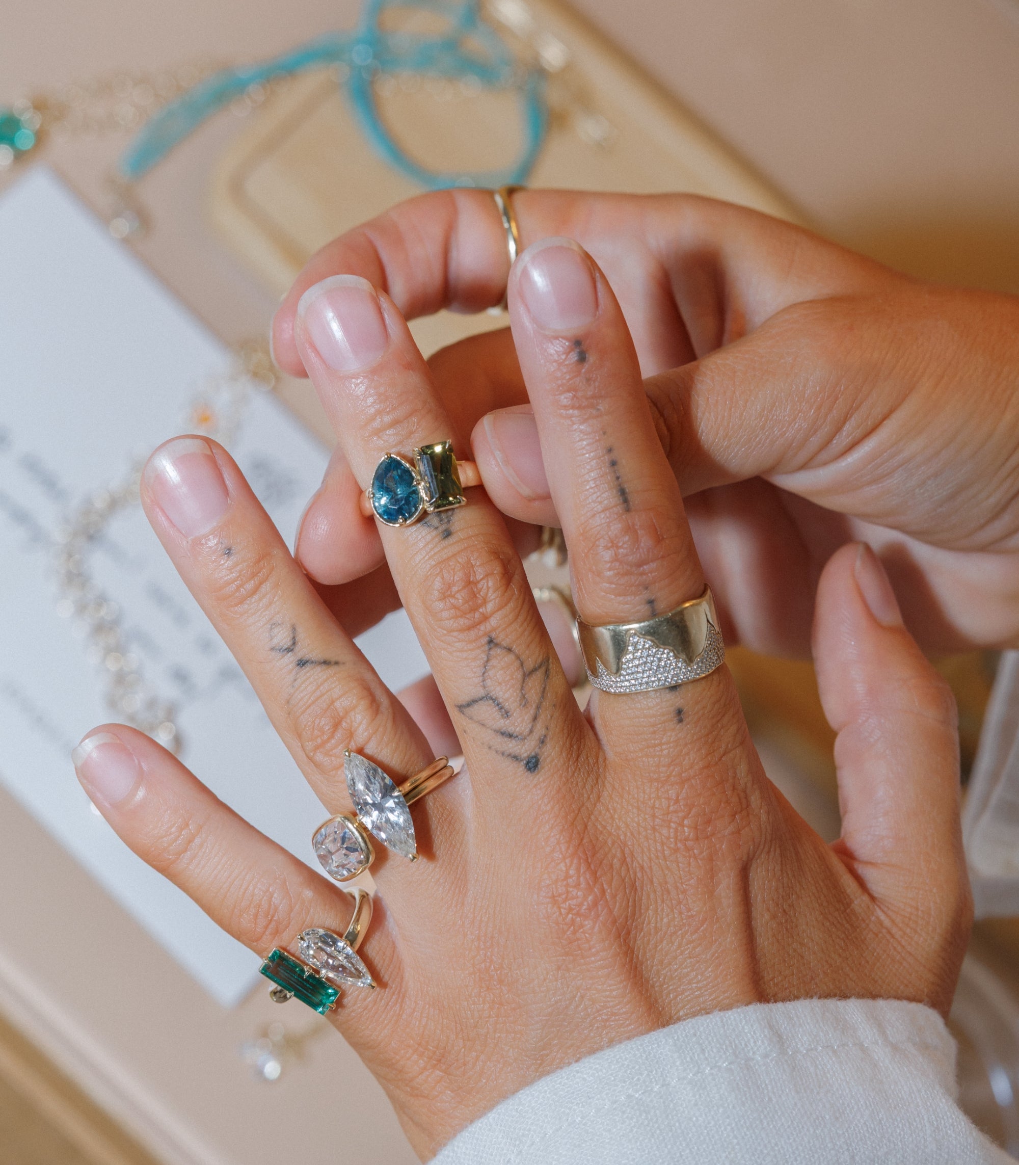 A close-up of a hand wearing multiple bohemian rings, including a textured silver band, a gold ring with a blue stone, and other silver rings with green and clear stones, all designed by Sofia Kaman  and would all make fabulous unique engagement rings.