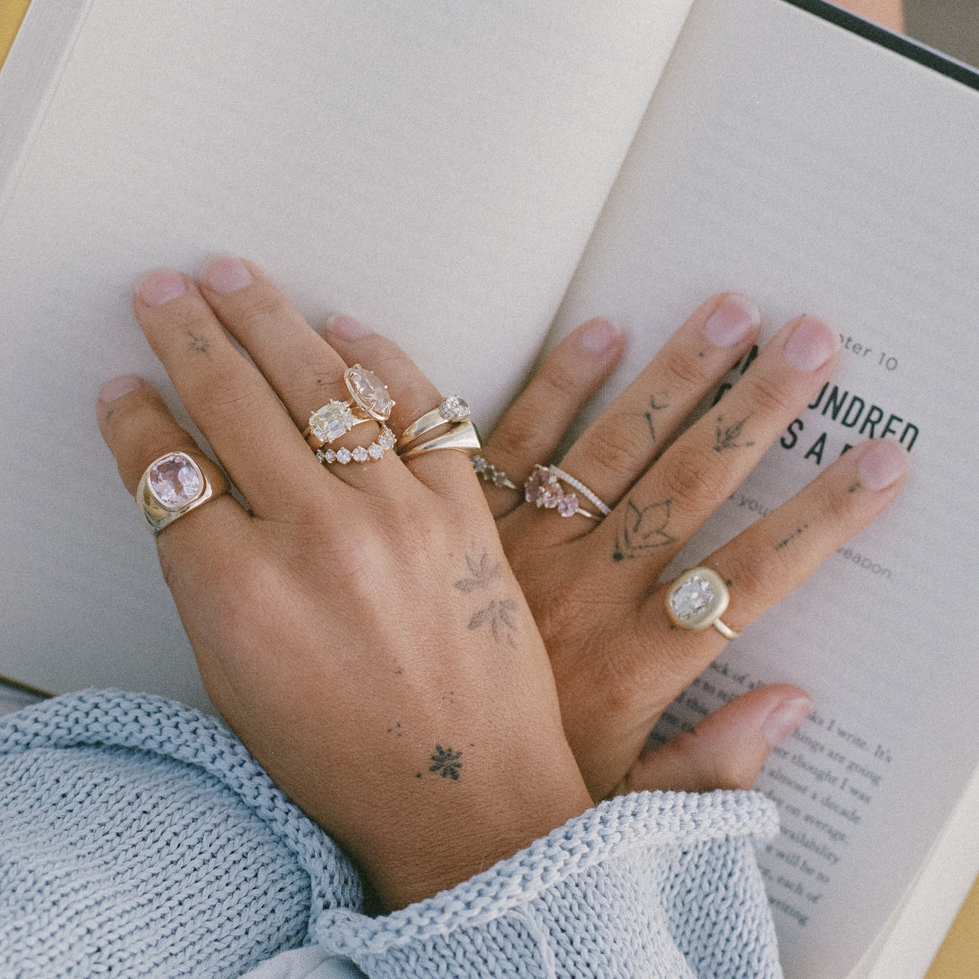 A close-up of two hands resting on an open book, wearing multiple gold rings with clear gemstones. The hands have small floral tattoos, and the person is wearing a light blue knit sweater.